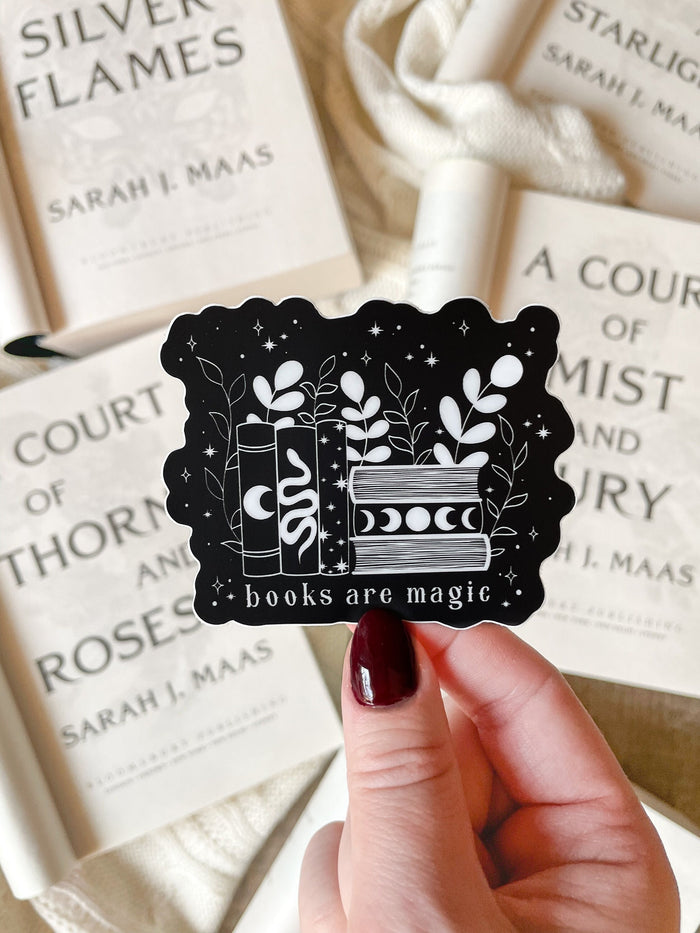 This "Books Are Magic" waterproof vinyl sticker has a black background with a white design and text. The design is a small bookshelf with 6 books. The 3 on the left are vertical with a moon, serpent, and stars on them. The three on the right are stacked horizontally with the top and bottom one showing the pages and the middle ones spine has a moon phase image. The books are surrounded by foliage and little stars.