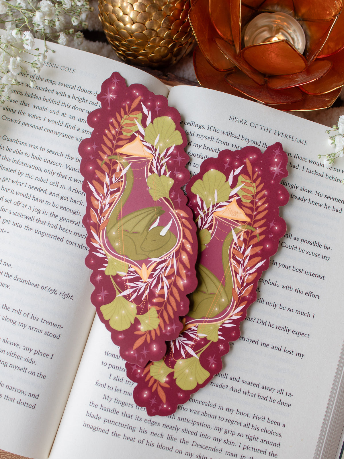 This fantasy autumnal fall sleeping dragon elixir bookmark features a little green dragon curled up in a potion bottle with their tale sticking out the top. Surrounded by orange, green, and light pink foliage with little stars and magic bubbles floating around. On a maroon background