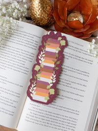 This autumn fall bookshelf bookmark has a maroon background and a stack of warm tones books in a lighter maroon, orange, rust, and light pink. Surrounded by foliage in various shades of green and little stars.