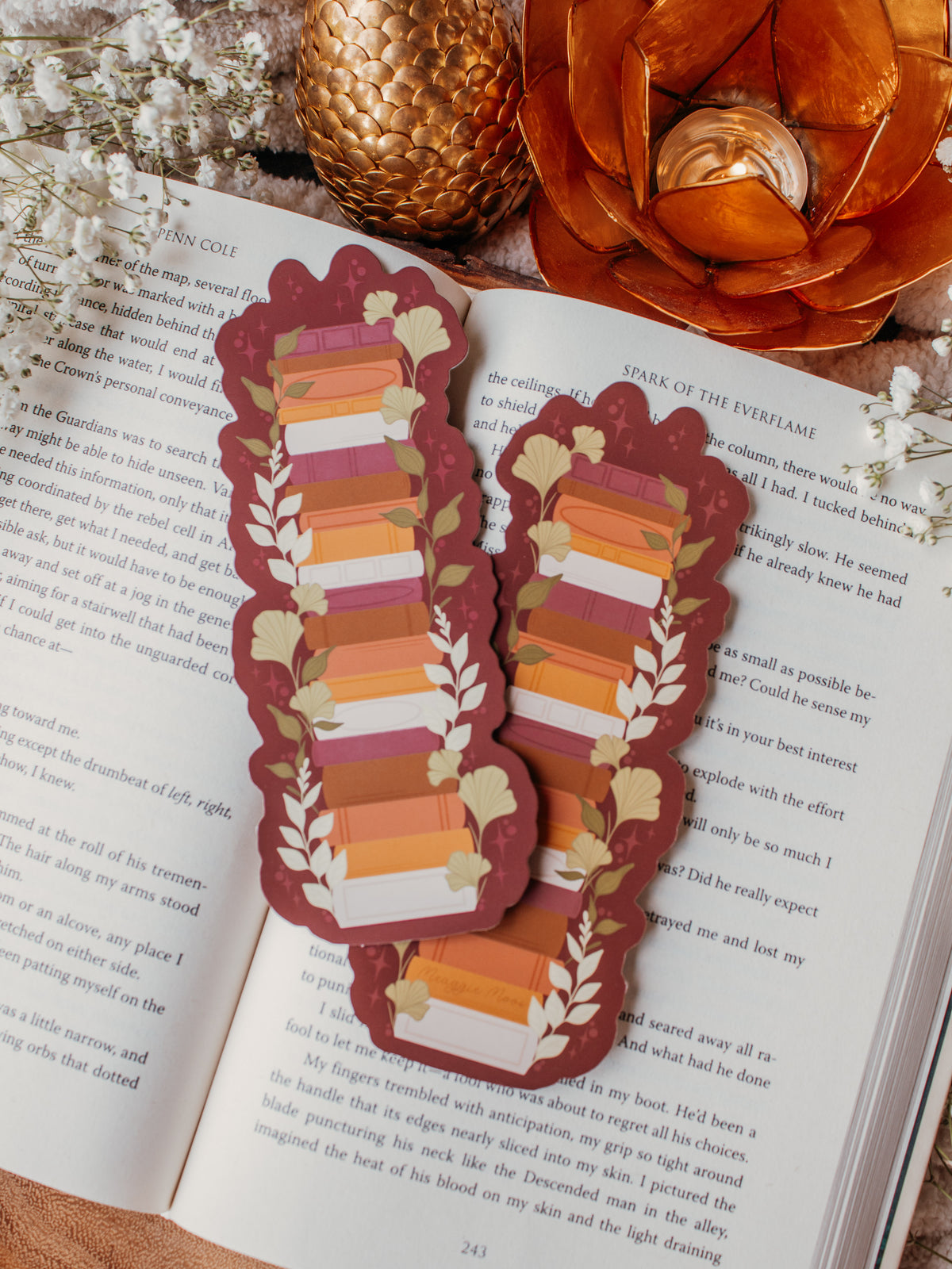 This autumn fall bookshelf bookmark has a maroon background and a stack of warm tones books in a lighter maroon, orange, rust, and light pink. Surrounded by foliage in various shades of green and little stars.