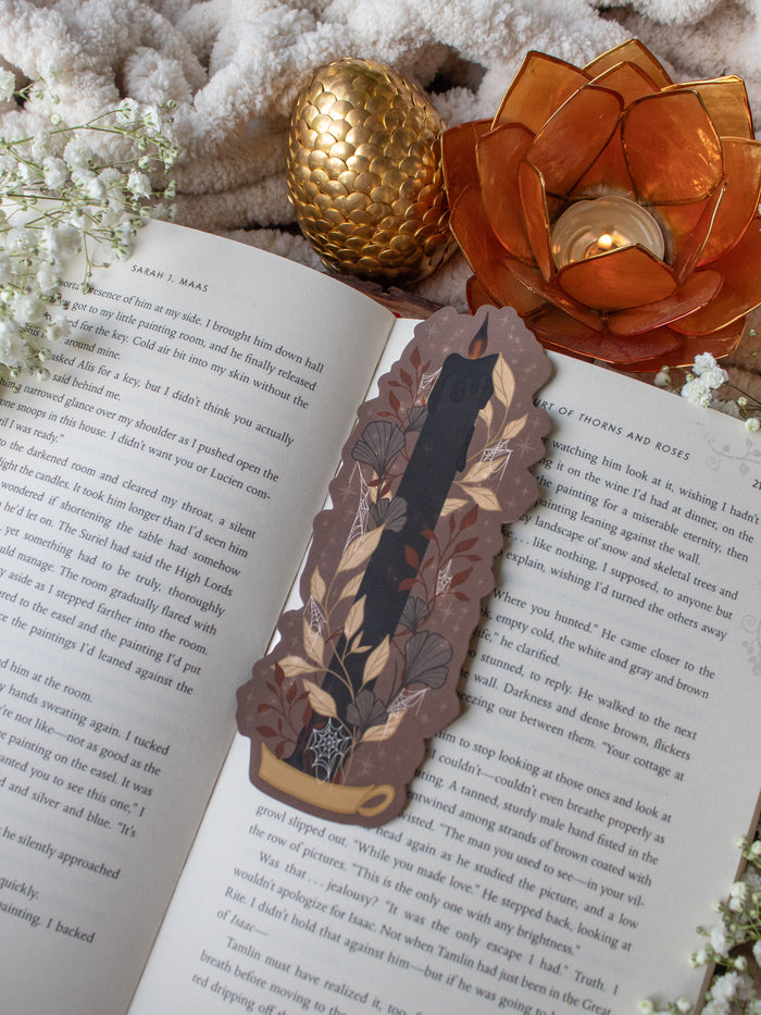 This spooky Halloween Black Flame candle bookmark is a black candlestick in a holder on a dark taupe background with light tan, burnt orange, and dark grey/ charcoal foliage surrounding it. The flame is tinged black and there are little stars and magic bubbles floating around.
