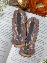 This spooky Halloween Black Flame candle bookmark is a black candlestick in a holder on a dark taupe background with light tan, burnt orange, and dark grey/ charcoal foliage surrounding it. The flame is tinged black and there are little stars and magic bubbles floating around.