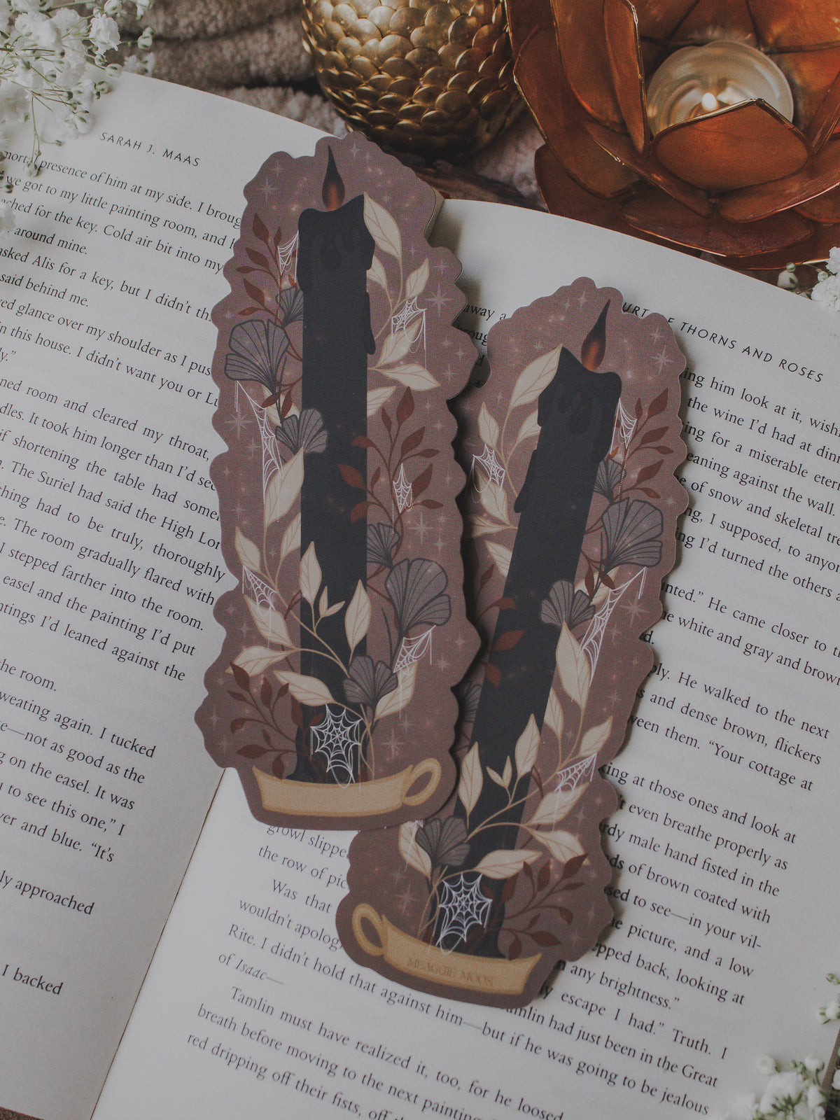 This spooky Halloween Black Flame candle bookmark is a black candlestick in a holder on a dark taupe background with light tan, burnt orange, and dark grey/ charcoal foliage surrounding it. The flame is tinged black and there are little stars and magic bubbles floating around.