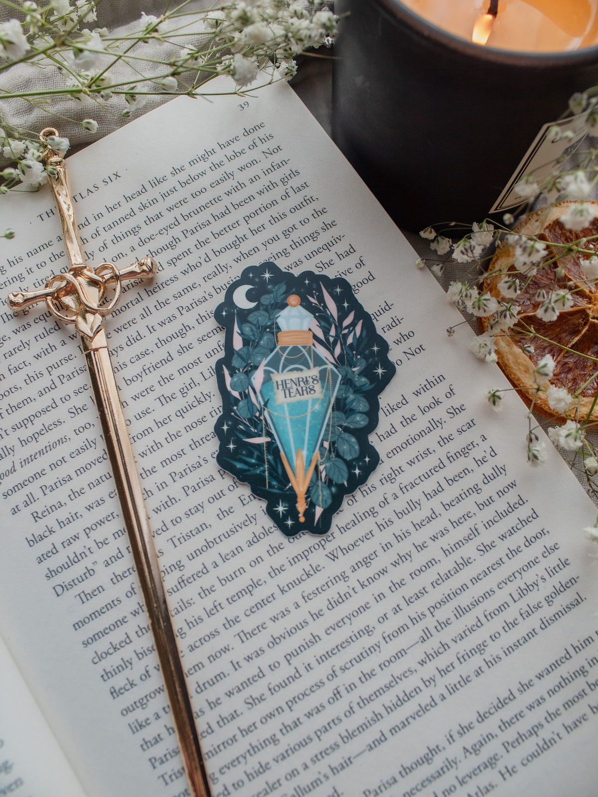 Spark Of The Everflame Henri's Tears Potion Sticker from the Kindred's Curse Saga by Penn Cole. This officially licensed sticker has a dark teal background, with a diamond shaped potion, light teal contents, surrounded by various shades of teal and pink foliage, stars, a crescent moon, and lil magical bubbles.
