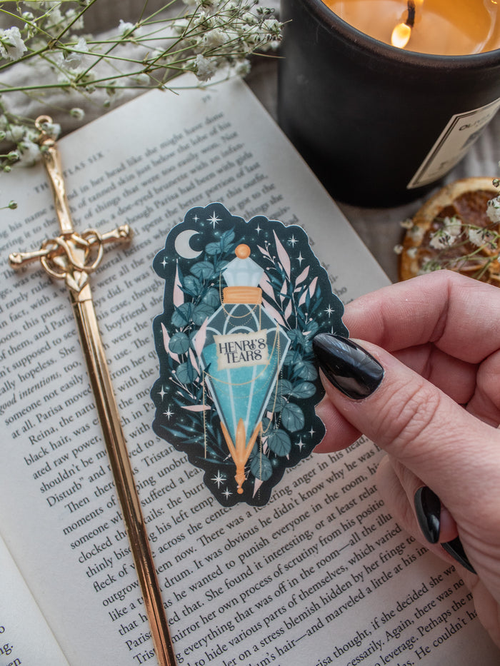 Spark Of The Everflame Henri's Tears Potion Sticker from the Kindred's Curse Saga by Penn Cole. This officially licensed sticker has a dark teal background, with a diamond shaped potion, light teal contents, surrounded by various shades of teal and pink foliage, stars, a crescent moon, and lil magical bubbles.