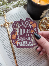 Maroon Sticker with the text "low So Bright The Darkness Trembles" from Spark of the Everflame by Penn Cole from the Kindred's Curse Saga. The text on this officially licensed sticker is surrounded by orange and tan foliage, stars, and magical looking bubbles.