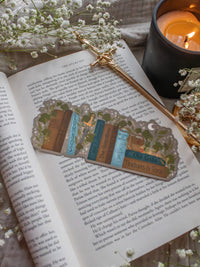 Inspired by Spark of the Everflame from the Kindreds Curse Sage by Penn Cole, this officially licensed Diem's bookshelf bookmark features a bookshelf in earth tones, dark teal blue and light sky blue. The books have titles like "The Guardians", Old Gods, Mortal Medicine and other nods at the series. Surrounded by foliage, the Sword of Corbois, a Lumnos healers clinic satchel, a moon shaped flameroot bottle, and Diem's Lunmos crown.
