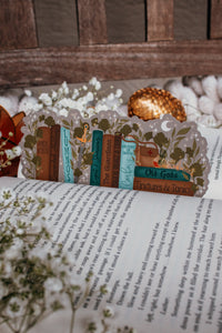 Inspired by Spark of the Everflame from the Kindreds Curse Sage by Penn Cole, this officially licensed Diem's bookshelf bookmark features a bookshelf in earth tones, dark teal blue and light sky blue. The books have titles like "The Guardians", Old Gods, Mortal Medicine and other nods at the series. Surrounded by foliage, the Sword of Corbois, a Lumnos healers clinic satchel, a moon shaped flameroot bottle, and Diem's Lunmos crown.