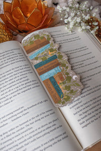 Inspired by Spark of the Everflame from the Kindreds Curse Sage by Penn Cole, this officially licensed Diem's bookshelf bookmark features a bookshelf in earth tones, dark teal blue and light sky blue. The books have titles like "The Guardians", Old Gods, Mortal Medicine and other nods at the series. Surrounded by foliage, the Sword of Corbois, a Lumnos healers clinic satchel, a moon shaped flameroot bottle, and Diem's Lunmos crown.