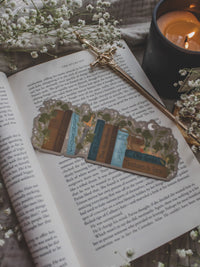 Inspired by Spark of the Everflame from the Kindreds Curse Sage by Penn Cole, this officially licensed Diem's bookshelf bookmark features a bookshelf in earth tones, dark teal blue and light sky blue. The books have titles like "The Guardians", Old Gods, Mortal Medicine and other nods at the series. Surrounded by foliage, the Sword of Corbois, a Lumnos healers clinic satchel, a moon shaped flameroot bottle, and Diem's Lunmos crown.