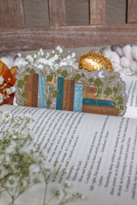Inspired by Spark of the Everflame from the Kindreds Curse Sage by Penn Cole, this officially licensed Diem's bookshelf bookmark features a bookshelf in earth tones, dark teal blue and light sky blue. The books have titles like "The Guardians", Old Gods, Mortal Medicine and other nods at the series. Surrounded by foliage, the Sword of Corbois, a Lumnos healers clinic satchel, a moon shaped flameroot bottle, and Diem's Lunmos crown.