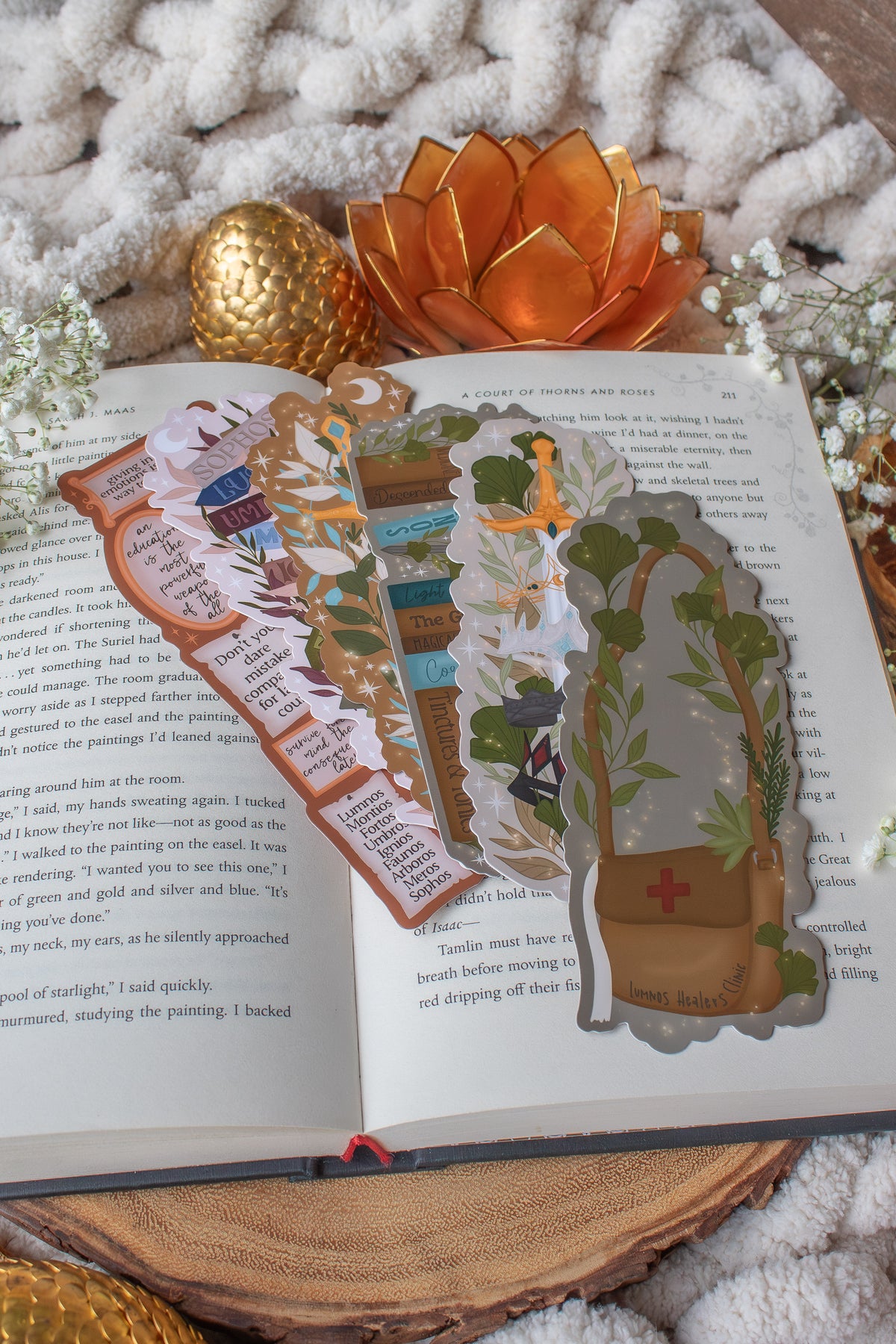 Inspired by Spark of the Everflame from the Kindreds Curse Sage by Penn Cole, this officially licensed Diem's bookshelf bookmark features a bookshelf in earth tones, dark teal blue and light sky blue. The books have titles like "The Guardians", Old Gods, Mortal Medicine and other nods at the series. Surrounded by foliage, the Sword of Corbois, a Lumnos healers clinic satchel, a moon shaped flameroot bottle, and Diem's Lunmos crown.