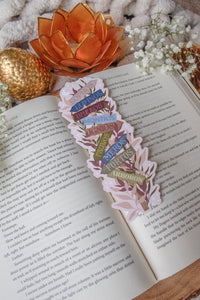This Spark of the Everflame Emarion Location Sign Bookmark has a wooden post with arrows pointing in different directions in various earth tone shades. The arrows are each labeled with one of the realms of Emarion. The bookmark has a light taupe background and there is foliage in pink and maroon surrounding the sign.