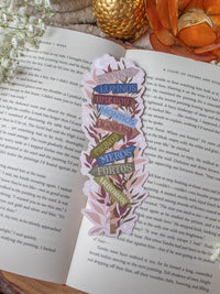 This Spark of the Everflame Emarion Location Sign Bookmark has a wooden post with arrows pointing in different directions in various earth tone shades. The arrows are each labeled with one of the realms of Emarion. The bookmark has a light taupe background and there is foliage in pink and maroon surrounding the sign.