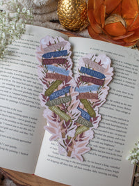This Spark of the Everflame Emarion Location Sign Bookmark has a wooden post with arrows pointing in different directions in various earth tone shades. The arrows are each labeled with one of the realms of Emarion. The bookmark has a light taupe background and there is foliage in pink and maroon surrounding the sign.