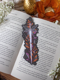 This spooky Halloween Haunted Faerie Dagger dagger bookmark is a long silver dagger with whispy faerie wings that look like spiderwebs. Surrounded but orange, green and maroon foliage covered in spiderwebs. Little stars and magic bubbles all around. Black background.