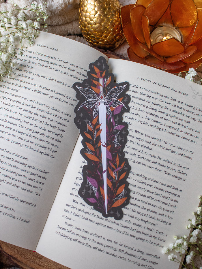 This spooky Halloween Haunted Faerie Dagger dagger bookmark is a long silver dagger with whispy faerie wings that look like spiderwebs. Surrounded but orange, green and maroon foliage covered in spiderwebs. Little stars and magic bubbles all around. Black background.