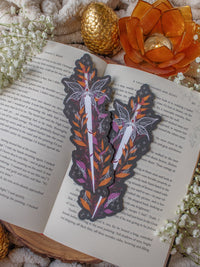 This spooky Halloween Haunted Faerie Dagger dagger bookmark is a long silver dagger with whispy faerie wings that look like spiderwebs. Surrounded but orange, green and maroon foliage covered in spiderwebs. Little stars and magic bubbles all around. Black background.