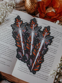 This spooky Halloween Haunted Faerie Dagger dagger bookmark is a long silver dagger with whispy faerie wings that look like spiderwebs. Surrounded but orange, green and maroon foliage covered in spiderwebs. Little stars and magic bubbles all around. Black background.