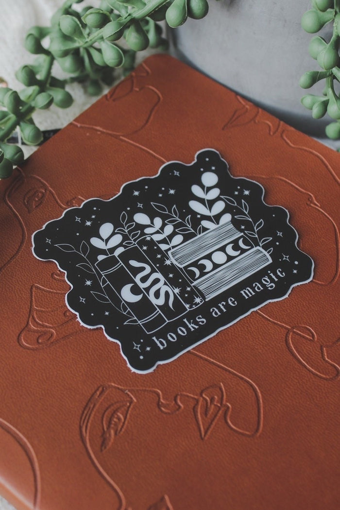 This "Books Are Magic" waterproof vinyl sticker has a black background with a white design and text. The design is a small bookshelf with 6 books. The 3 on the left are vertical with a moon, serpent, and stars on them. The three on the right are stacked horizontally with the top and bottom one showing the pages and the middle ones spine has a moon phase image. The books are surrounded by foliage and little stars.