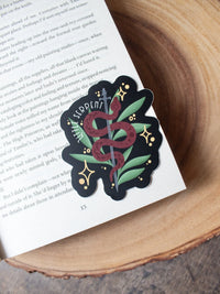 This Little Serpent sticker from The Serpent and The Wings of Night from the Crowns of Nyaxia Series by Carissa Broadbent is officially licensed. It features a maroon snake wrapped around a steel sword in front of green foliage on a black background with the text "Little Serpent" around the body of the snake.