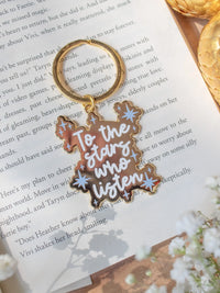 This officially licensed "To The Stars Who Listen" metal keychain is inspired from A Court of Thorns and Roses by Sarah J Maas. It is gold-colored, with the "To The Stars Who Listen" in a white bold script and little light blue stars around it.