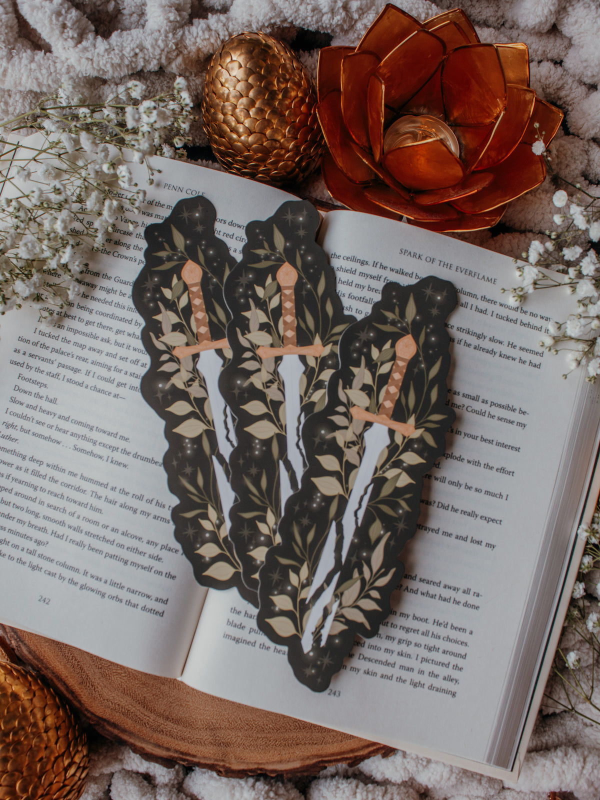 Broken Fantasy Sword Die-Cut Bookmark on an open book with a gold metal dragon egg, baby's breath, and an orange lotus tea light holder in the background. 