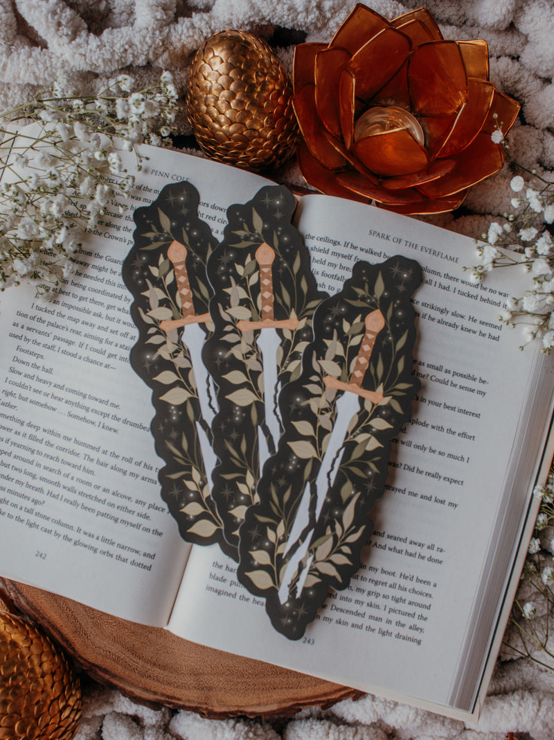 Broken Fantasy Sword Die-Cut Bookmark on an open book with a gold metal dragon egg, baby's breath, and an orange lotus tea light holder in the background. 