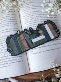 ACOTAR Funny Feyre Bookshelf Bookmark from A Court of Thorns and Roses by Sarah J Maas. A Bookshelf in earth tones like deep greens, browns and charcoal with a cauldron and a "snowball fight champion" trophy with book titles like "Reasons Tamlin is a lil bitch", "A Gardeners guide to strictly inedible plants", "Prison Breaks 101" and more.