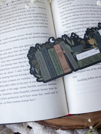 ACOTAR Funny Feyre Bookshelf Bookmark from A Court of Thorns and Roses by Sarah J Maas. A Bookshelf in earth tones like deep greens, browns and charcoal with a cauldron and a "snowball fight champion" trophy with book titles like "Reasons Tamlin is a lil bitch", "A Gardeners guide to strictly inedible plants", "Prison Breaks 101" and more.