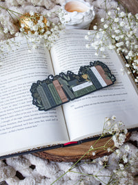 ACOTAR Funny Feyre Bookshelf Bookmark from A Court of Thorns and Roses by Sarah J Maas. A Bookshelf in earth tones like deep greens, browns and charcoal with a cauldron and a "snowball fight champion" trophy with book titles like "Reasons Tamlin is a lil bitch", "A Gardeners guide to strictly inedible plants", "Prison Breaks 101" and more.