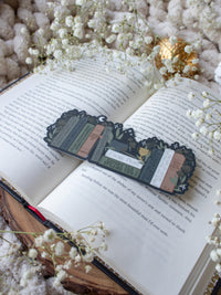 ACOTAR Funny Feyre Bookshelf Bookmark from A Court of Thorns and Roses by Sarah J Maas. A Bookshelf in earth tones like deep greens, browns and charcoal with a cauldron and a "snowball fight champion" trophy with book titles like "Reasons Tamlin is a lil bitch", "A Gardeners guide to strictly inedible plants", "Prison Breaks 101" and more.