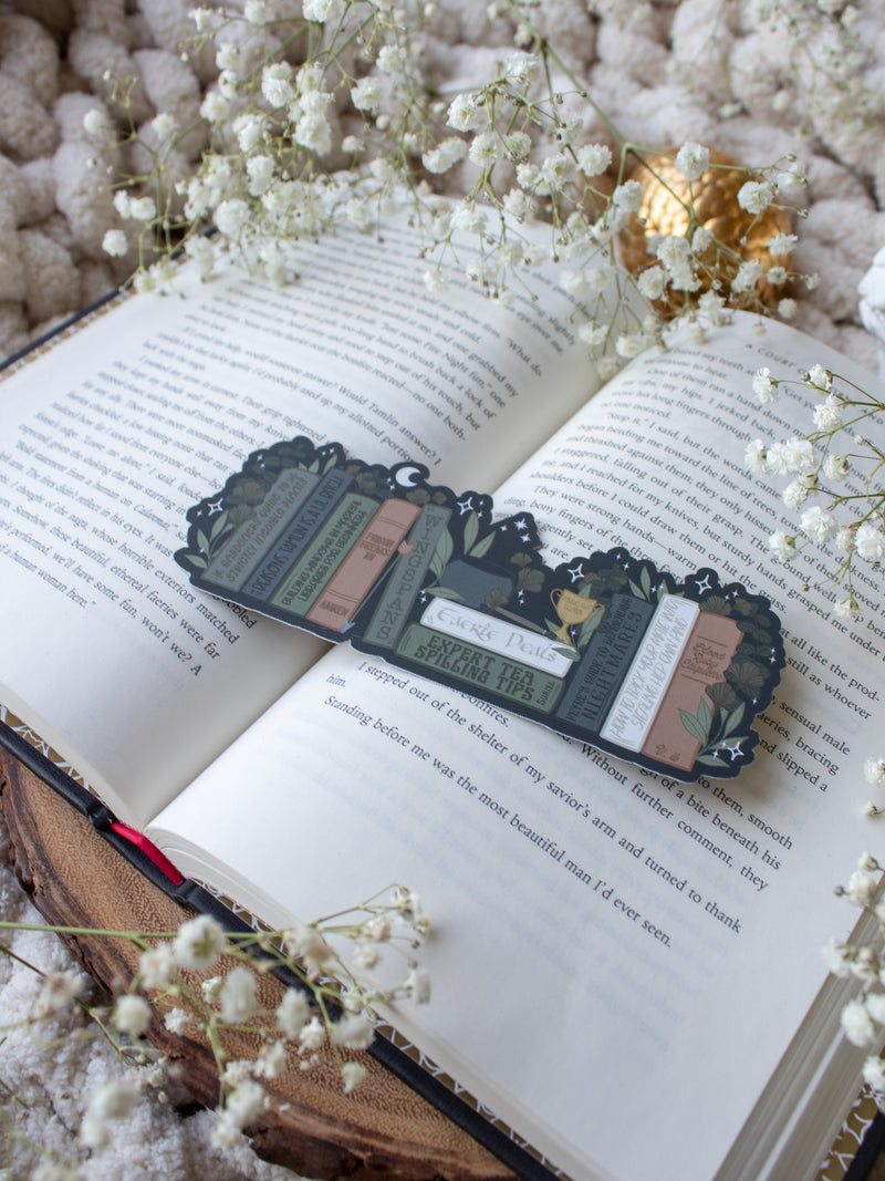 ACOTAR Funny Feyre Bookshelf Bookmark from A Court of Thorns and Roses by Sarah J Maas. A Bookshelf in earth tones like deep greens, browns and charcoal with a cauldron and a "snowball fight champion" trophy with book titles like "Reasons Tamlin is a lil bitch", "A Gardeners guide to strictly inedible plants", "Prison Breaks 101" and more.