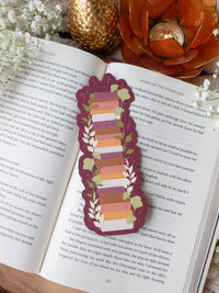 This autumn fall bookshelf bookmark has a maroon background and a stack of warm tones books in a lighter maroon, orange, rust, and light pink. Surrounded by foliage in various shades of green and little stars. 