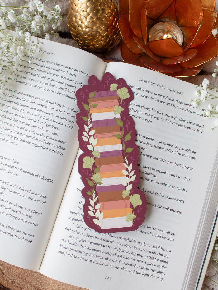 This autumn fall bookshelf bookmark has a maroon background and a stack of warm tones books in a lighter maroon, orange, rust, and light pink. Surrounded by foliage in various shades of green and little stars. 