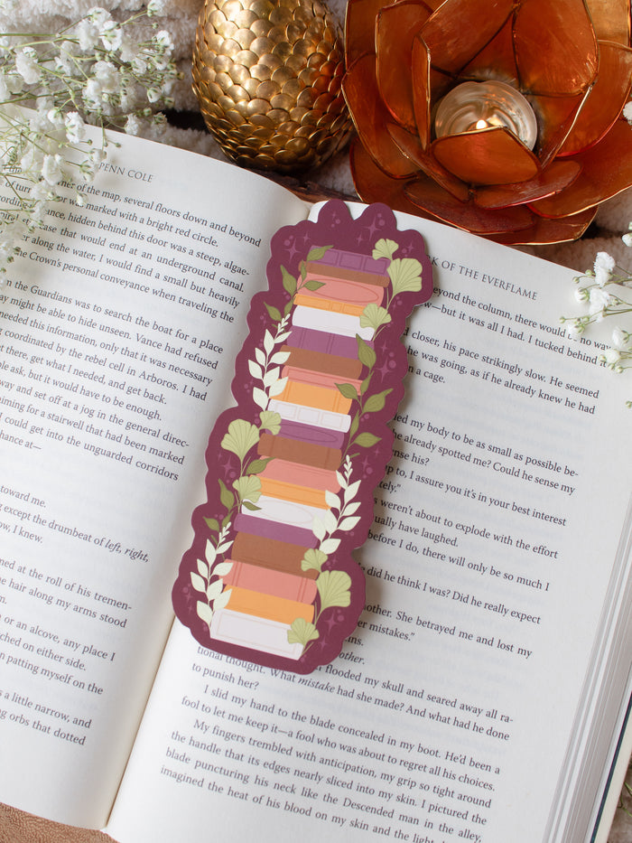 This autumn fall bookshelf bookmark has a maroon background and a stack of warm tones books in a lighter maroon, orange, rust, and light pink. Surrounded by foliage in various shades of green and little stars. 