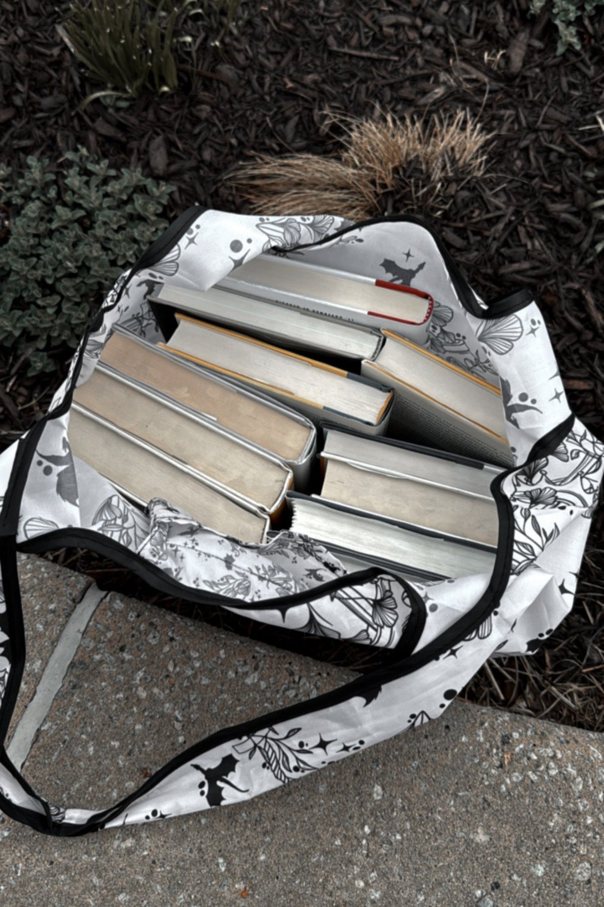 A fantasy reader dragon covered reusable tote bag set against a ledge and filled with tons of books. 