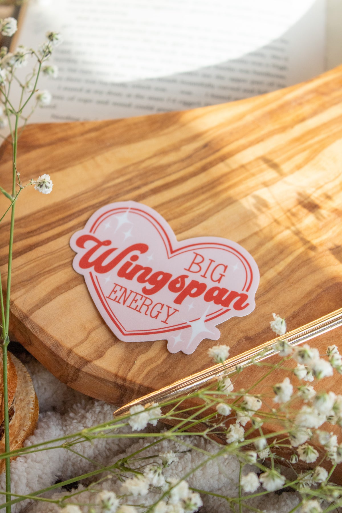 Big Wingspan Energy Romantasy Reader Waterproof Vinyl Sticker 

A pink heart  sticker with red text saying "Big Wingspan Energy" and white stars around it. Shown on a wood cutting board on a book with little white flowers around it. 