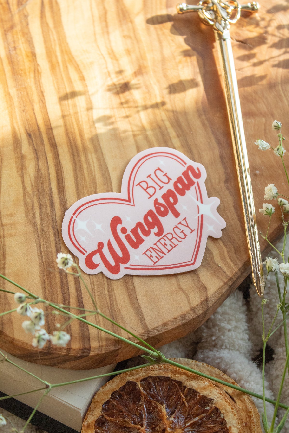 Big Wingspan Energy Romantasy Reader Waterproof Vinyl Sticker 

A pink heart  sticker with red text saying "Big Wingspan Energy" and white stars around it. Shown on a wood cutting board on a book with little white flowers around it. 