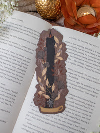 This spooky Halloween Black Flame candle bookmark is a black candlestick in a holder on a dark taupe background with light tan, burnt orange, and dark grey/ charcoal foliage surrounding it. The flame is tinged black and there are little stars and magic bubbles floating around. 
