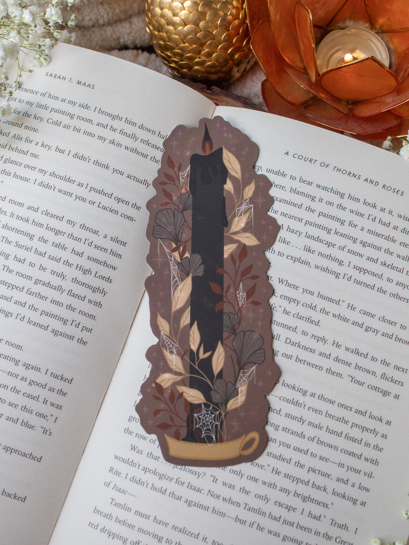 This spooky Halloween Black Flame candle bookmark is a black candlestick in a holder on a dark taupe background with light tan, burnt orange, and dark grey/ charcoal foliage surrounding it. The flame is tinged black and there are little stars and magic bubbles floating around. 