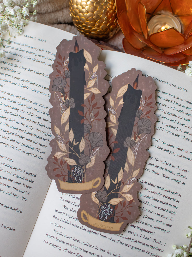 This spooky Halloween Black Flame candle bookmark is a black candlestick in a holder on a dark taupe background with light tan, burnt orange, and dark grey/ charcoal foliage surrounding it. The flame is tinged black and there are little stars and magic bubbles floating around. 