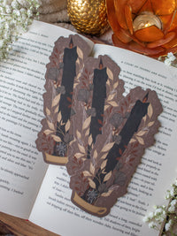 This spooky Halloween Black Flame candle bookmark is a black candlestick in a holder on a dark taupe background with light tan, burnt orange, and dark grey/ charcoal foliage surrounding it. The flame is tinged black and there are little stars and magic bubbles floating around. 