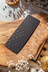This Black and Gold Dragon Scale Foil bookmark for Fantasy and Romantasy Readers is rectangular in shape and has a black background with gold foil scales. 

Shown on an olive wood cutting board, on an open book sitting on a chunky knit blanket with baby's Breath flowers, orange slices, and gold dragon eggs surrounding it. 