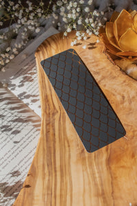 This Black and Gold Dragon Scale Foil bookmark for Fantasy and Romantasy Readers is rectangular in shape and has a black background with gold foil scales. 

Shown on an olive wood cutting board, on an open book sitting on a chunky knit blanket with baby's Breath flowers, orange slices, and gold dragon eggs surrounding it. 