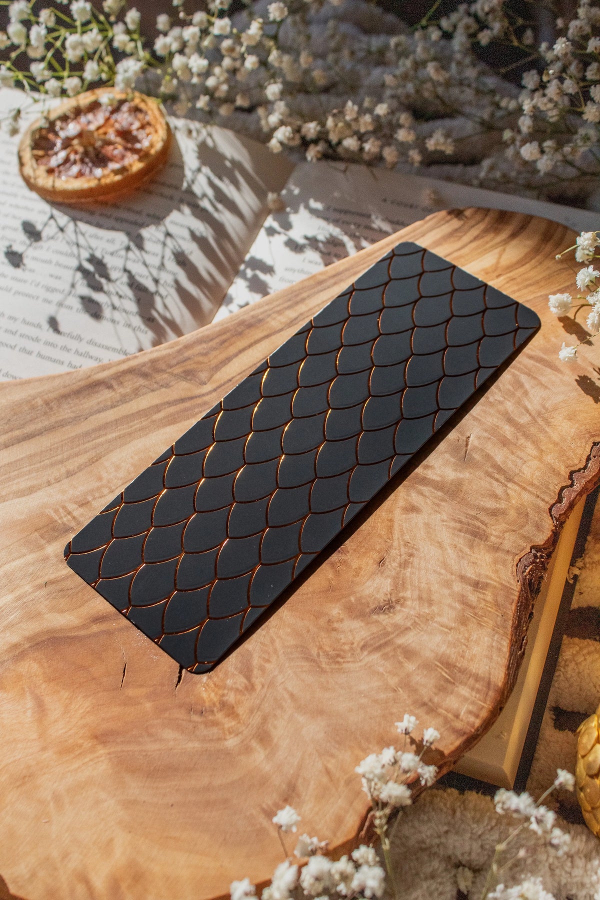 This Black and Gold Dragon Scale Foil bookmark for Fantasy and Romantasy Readers is rectangular in shape and has a black background with gold foil scales. 

Shown on an olive wood cutting board, on an open book sitting on a chunky knit blanket with baby's Breath flowers, orange slices, and gold dragon eggs surrounding it. 