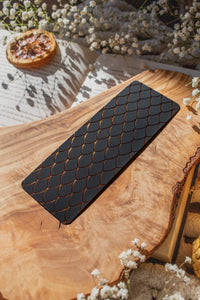 This Black and Gold Dragon Scale Foil bookmark for Fantasy and Romantasy Readers is rectangular in shape and has a black background with gold foil scales. 

Shown on an olive wood cutting board, on an open book sitting on a chunky knit blanket with baby's Breath flowers, orange slices, and gold dragon eggs surrounding it. 