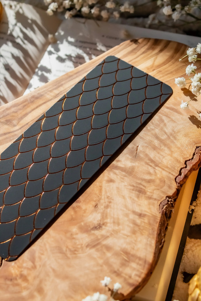 This Black and Gold Dragon Scale Foil bookmark for Fantasy and Romantasy Readers is rectangular in shape and has a black background with gold foil scales. 

Shown on an olive wood cutting board, on an open book sitting on a chunky knit blanket with baby's Breath flowers, orange slices, and gold dragon eggs surrounding it. 