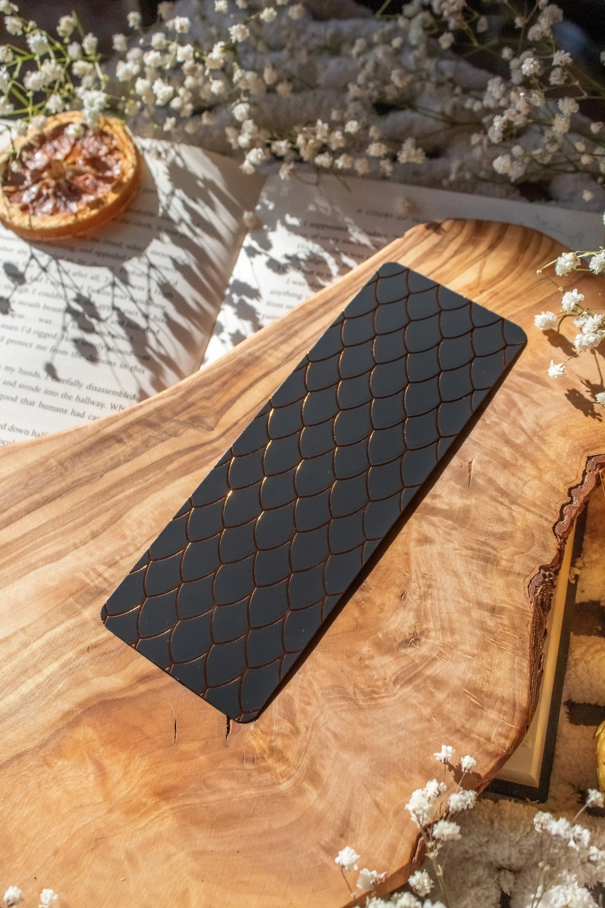 This Black and Gold Dragon Scale Foil bookmark for Fantasy and Romantasy Readers is rectangular in shape and has a black background with gold foil scales. 

Shown on an olive wood cutting board, on an open book sitting on a chunky knit blanket with baby's Breath flowers, orange slices, and gold dragon eggs surrounding it. 