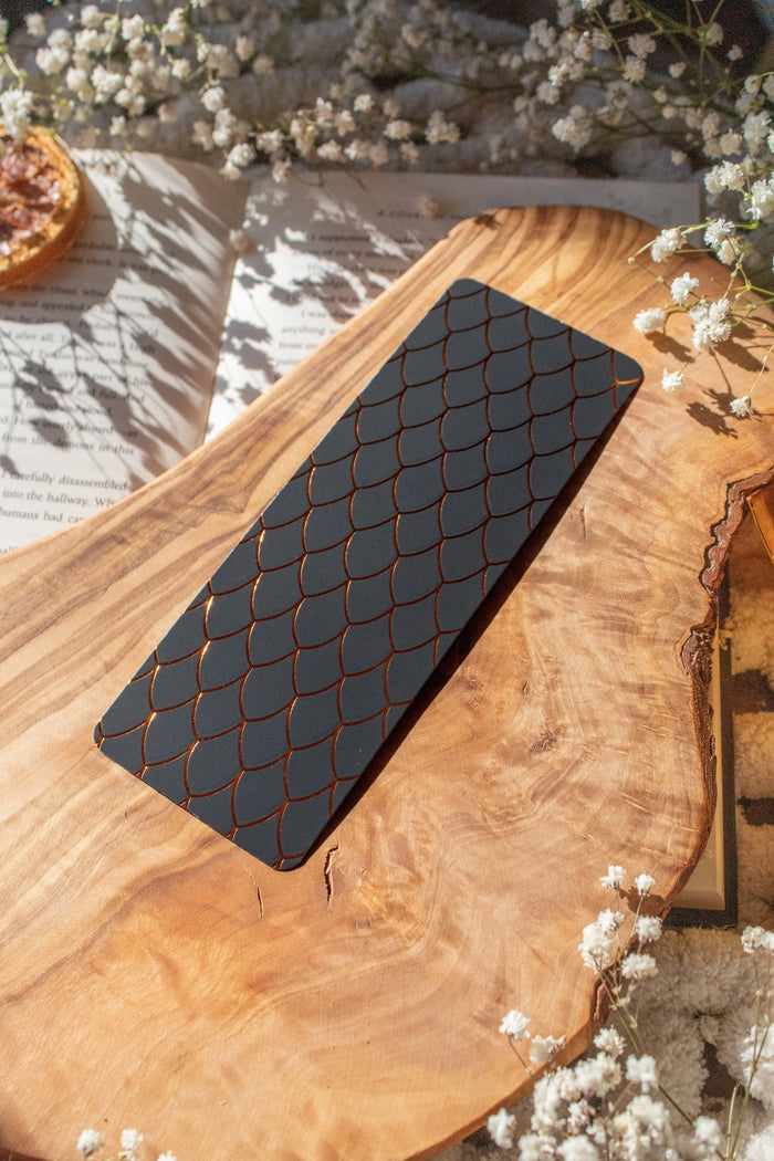 This Black and Gold Dragon Scale Foil bookmark for Fantasy and Romantasy Readers is rectangular in shape and has a black background with gold foil scales. 

Shown on an olive wood cutting board, on an open book sitting on a chunky knit blanket with baby's Breath flowers, orange slices, and gold dragon eggs surrounding it. 